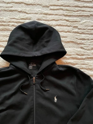 Svart hoodie med dragkedja Polo Ralph Lauren - Svart hoodie med dragkedja från Polo Ralph Lauren. Klassisk design med huva och dragsko, två fickor framtill och broderad logga på bröstet. Tillverkad i mjukt bomullstyg för en skön och avslappnad stil. Mycket bra skick, endast lite nopprig som ska gå bort med en noppmaskin Längd 69cm. Bredd: 58cm