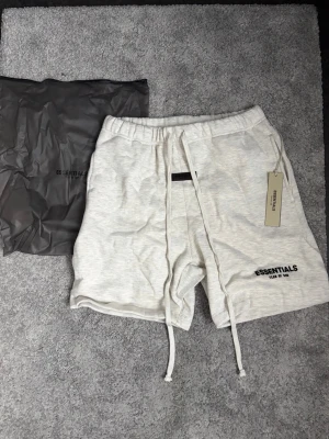 EssentialsShorts - Snygga ljusgrå shorts med långa snören i midjan och broderad logga på benet. Mjukt material och elastisk midja gör dem riktigt bekväma. Perfekta för chill dagar eller när du vill ha en clean look.