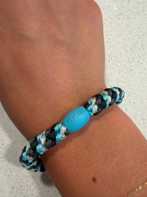 Kknekki hårband - Snyggt flätat armband i blå, svart och vit nylon med en blå plastdetalj där det står 'Kknekki'. Perfekt accessoar för en cool och avslappnad stil. Armbandet är elastiskt och passar de flesta handleder.