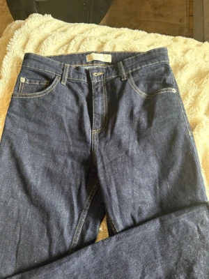 Mörkblå raka jeans i denim - Säljer ett par klassiska mörkblå jeans i rå denim med raka ben och kontrastsömmar. Jeansen har fem fickor, bälteshällor och stängs med knapp och dragkedja. Ny med prislapp då jag glömt returnera dem 