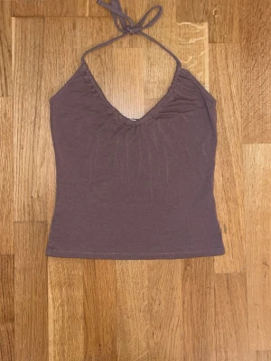 Brun halterneck linnetopp - Säljer en brun halterneck linnetopp med knyt i nacken och v-ringad front. Toppen är ärmlös och har en enkel, stilren design i mjukt bomullsmaterial. Perfekt för varma dagar eller att styla lager på lager.