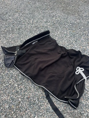 Svart fleecetäcke för häst - Säljer ett svart fleecetäcke för häst med vita kantband och dekorativt snöre. Täcket har spänne fram och remmar under magen. Perfekt för ridning eller transport.