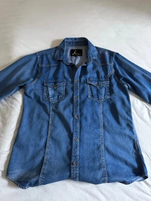 Jeansskjorta  - Snygg blå jeansskjorta från Qandil med klassisk krage, knappar framtill och två bröstfickor med lock. Skjortan har lång ärm. Perfekt för en avslappnad och cool stil.