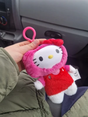 Hello Kitty mjukisdjur med vinylansikte - Söt Hello Kitty mjukisdjur med vinylansikte, klädd i röd klänning och rosa huva. Nyckelringsfäste i plast gör den enkel att hänga på väskan. Kommer i originalförpackning med färgglad design och texten 'Hello Kitty and Friends'. Perfekt för samlare eller som present.