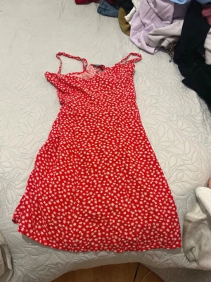 Röd blommig sommarklänning - Söt röd klänning med vita blommor och smala axelband. Kort modell med lätt utställd passform, perfekt för varma dagar. Tunt och luftigt material som känns bekvämt hela dagen.