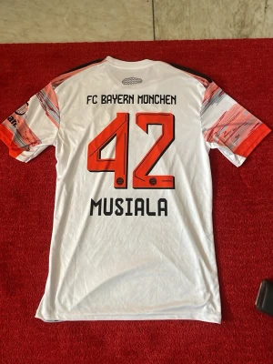 Bayern München Musiala matchtröja - Säljer en FC Bayern München matchtröja med Musiala och nummer 42 på ryggen. Tröjan är vit med röda och gråa grafiska detaljer, korta ärmar och klubbmärke på bröstet. Tillverkad i lätt och ventilerande material, perfekt för fotboll eller träning.