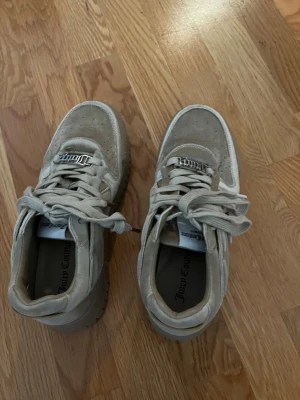 Beige sneakers Juicy Couture - Snygga beige sneakers från Juicy Couture med chunky sula och perforerade detaljer på tån. Skorna har breda snören och metallbricka med Juicy-logga framtill. Tillverkade i mockaliknande material för en trendig och sportig look.