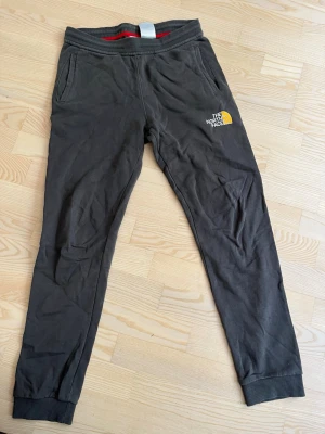 Svarta mjukisbyxor The North Face - Snygga svarta mjukisbyxor från The North Face med logga i vitt och gult på vänster ben. Byxorna har resår i midjan, ribbade muddar vid bensluten och två sidofickor. Perfekta för chill eller träning.