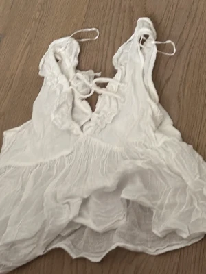 Vit linnetopp med snörning - Säljer en luftig vit linnetopp med tunna axelband och snygg snörning framtill. Toppen har volangdetaljer vid ringningen och är tillverkad i ett lätt, skirt material som passar perfekt till varma dagar..