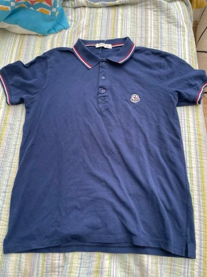 Marinblå pikétröja från Moncler - Snygg marinblå pikétröja från Moncler med klassisk krage och knappar framtill. Tröjan har vita och röda detaljer på kragen och ärmsluten samt Moncler-logga broderad på bröstet. Perfekt för en stilren och avslappnad look.