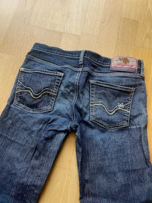 Mörkblå bootcut Replay jeans med brodyr - Säljer ett par mörkblå bootcut jeans med snygga kontrastsömmar och broderade fickor bak. Jeansen har klassisk femficksdesign och en patch med logga i midjan. Perfekta till en avslappnad stil och tillverkade i slitstarkt denimtyg. Jätte bekväm.