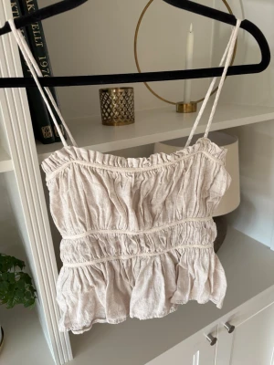 Beige volangtop med smala axelband - Supersöt beige topp med smala axelband och volangdetaljer. Toppen har en rynkad design och är gjord i ett luftigt linnematerial, perfekt för varma dagar. Passar dig som gillar en romantisk och avslappnad stil.