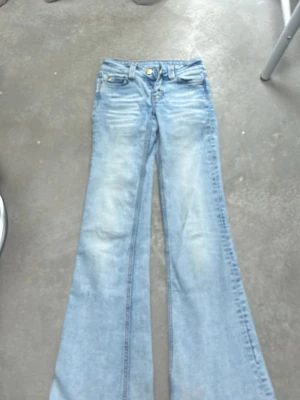 Ljusblå bootcut jeans med utsvängda ben - Säljer ett par ljusblå bootcut jeans med utsvängda ben och klassisk femficksmodell. Jeansen har slitningar framtill för en cool vintagekänsla och en grön patch bak i midjan. Perfekta för dig som gillar retrostil och vill sticka ut.