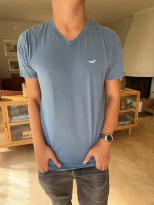 Blå v-ringad t-shirt Hollister - Snygg blå t-shirt från Hollister med v-ringning och diskret logga på bröstet. T-shirten har korta ärmar och är gjord i ett mjukt och lätt material som känns skönt mot huden. Perfekt till jeans eller shorts för en avslappnad look.
