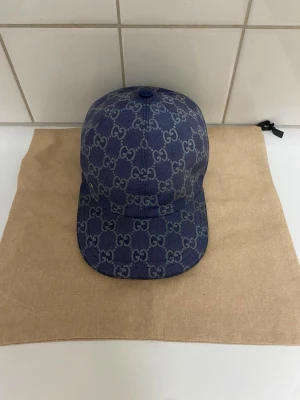 Blå Gucci keps med GG-mönster - Säljer en blå keps från Gucci med det klassiska GG-monogrammet över hela kepsen. Kepsen har böjd skärm och är tillverkad i ett denimliknande material. Perfekt för dig som vill ha en stilren och exklusiv accessoar till din outfit.