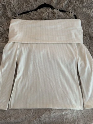 Vit ribbad offshoulder topp - Säljer en stilren cremevit offshoulder topp med ribbad struktur. Toppen har långa ärmar och en bred kant upptill som ger en snygg och trendig look. 