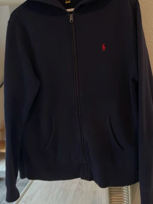Mörkblå hoodie från Polo Ralph Lauren - Snygg mörkblå hoodie från Polo Ralph Lauren med dragkedja framtill och klassisk röd logga på bröstet. Hoodien har huva, två fickor och är tillverkad i mjukt material som känns skönt mot huden. Perfekt för en avslappnad och stilren look. Storlek XL i ungdomsstorlek. Passar 176-180 ca. 