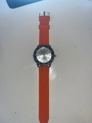 Armklocka från Quartz - Säljer en använd orange klocka från Quartz med rund silverfärgad urtavla, svart vridring och oranget armband. Klockan är använd ett par ggr men har inga tydliga tecken på använd. Perfekt accessoar för sig som vill sticka ut lite.