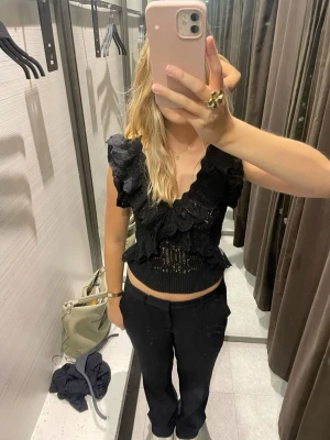 Volang topp från Zara - Säljer en svart blus med djup v-ringning, spetsdetaljer och volanger längs axlar och midja. Toppen är croppad och har ärmlös design med snygg struktur och romantisk känsla. Perfekt för dig som vill sticka ut lite extra.