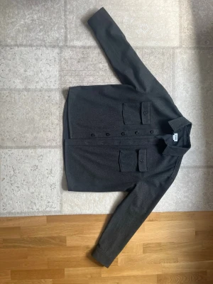 Mörkgrå overshirt med fickor - Stilren mörkgrå overshirt med raka linjer och två stora bröstfickor med lock och knappar. Klassisk krage och knäppning framtill. Materialet ser ut att vara tjockare, perfekt för lager på lager. Passar till många olika stilar.