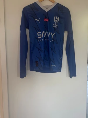 Blå långärmad Al Hilal matchtröja Puma - Snygg blå långärmad matchtröja från Al Hilal med vita och mörkblå blixtmönster. Tröjan har vit V-ringning, tryckta loggor från Puma, Jahez och Savvy Games Group samt DRYCELL-material som andas. Perfekt för fotbollsträning eller att bära till vardags.