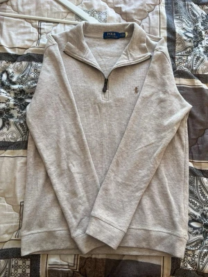 Beige quarter zip från Polo Ralph Lauren - Beige långärmad tröja från Polo Ralph Lauren med quarter zip och klassisk broderad logga på bröstet. Tillverkad i mjukt bomullsmaterial och har ribbade muddar vid ärmslut och nederkant. Perfekt för en avslappnad men stilren look.