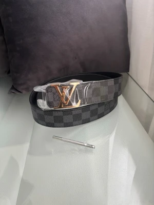 Bälte  - Snyggt svart bälte  med klassiskt rutigt mönster och stor logga i guld på spännet. Bältet har exklusiv känsla och passar perfekt till jeans eller kostymbyxor. Tillverkad i skinn och levereras med verktyg för justering.