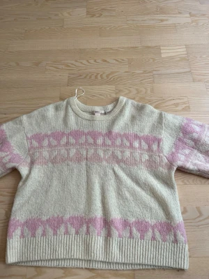 Vit stickad tröja med rosa mönster - Mysig stickad tröja i offwhite med breda rosa mönsterband över bröstet, ärmarna och nederkanten. Tröjan har rund halsringning och ribbstickade muddar. Perfekt för kalla dagar och ger en söt retrovibe.