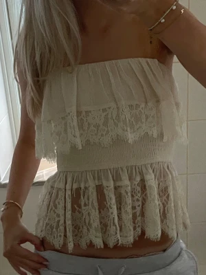 Beige spets tubtopp med volanger - Romantisk beige tubtopp med volanger och spetsdetaljer. Toppen är axelbandslös och har en smockad midja som ger en feminin siluett. Perfekt för dig som gillar en bohemisk och söt stil.
