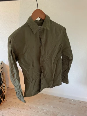 Olivgrön skjorta med knappar - Snygg olivgrön skjorta i bomull med klassisk krage och långa ärmar. Skjortan har knappar framtill och en enkel, stilren design som passar till mycket. Perfekt för dig som gillar en avslappnad men ändå clean look.