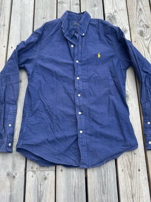 Marinblå skjorta från Polo Ralph Lauren - Klassisk marinblå skjorta från Polo Ralph Lauren med button-down krage och vit knappstängning. Skjortan har det ikoniska gula broderade logotypen på bröstet och är tillverkad i bomull. Perfekt för en stilren och avslappnad look.