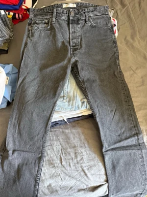 Grå jeans från Jack & Jones - Säljer ett par grå jeans från Jack & Jones med klassisk femficksmodell och raka ben. Jeansen har knappgylf och är tillverkade i bomull med en lätt stretch för extra komfort. Perfekta för en avslappnad och stilren look.