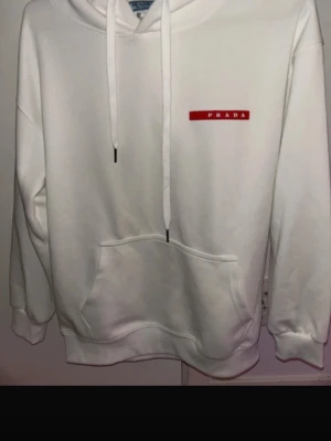Prada hoodie  - Snygg vit hoodie från Prada med klassisk röd logga på bröstet. Hoodien har huva med dragsko och en stor magficka framtill. Tillverkad i mjukt material som känns skönt mot huden. Perfekt för dig som vill ha en clean och stilren look.