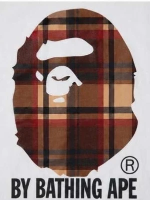 Vit BAPE t-shirt med rutig apa - Snygg vit t-shirt från BAPE med det klassiska apansiktet i rödbrunt rutigt mönster på bröstet och texten 'BY BATHING APE' under. Perfekt för dig som gillar streetwear och vill sticka ut med en unik design.