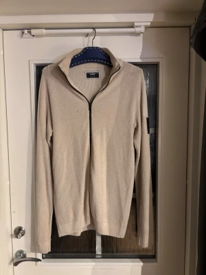 Beige ziptröja från Jack & Jones - Snygg beige ziptröja från Jack & Jones med hög krage och svart dragkedja. Tröjan har ett diskret märke på ärmen och är tillverkad i ett stickat material med struktur. Perfekt för lager på lager och enkel att matcha med olika outfits.