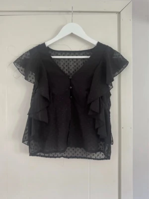 Svart blus med volanger - Snygg svart blus. Blusen har v-ringning, knäppning fram och korta ärmar. Perfekt för en trendig och luftig look.