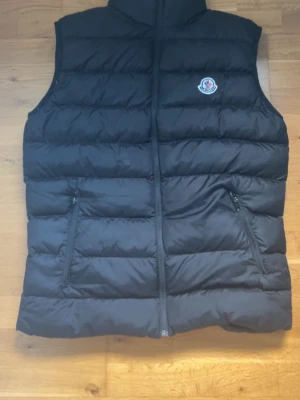 Svart dunväst från Moncler - Snygg svart dunväst från Moncler med klassisk quiltad design och hög krage. Västen har två dragkedjeförsedda fickor och Moncler-logga på bröstet. Perfekt för lager-på-lager och ger en clean look till din outfit. Den har en söndrig ficka på vänster sida men annars är det inget fel på den