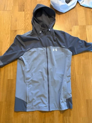 Grå vindjacka från Under Armour - Snygg grå vindjacka från Under Armour med dragkedja framtill och huva. Jackan har två olika grå nyanser och en vit logga på bröstet. Perfekt för blåsiga dagar och sportiga outfits.