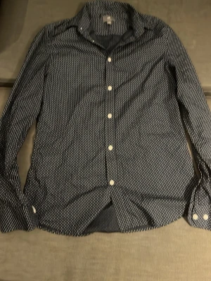 Mörkblå prickig skjorta H&M Slim Fit - Snygg mörkblå skjorta från H&M i slim fit-modell med vita prickar och klassisk krage. Skjortan har långa ärmar och vita knappar framtill. Perfekt för dig som gillar stilrena detaljer och enkel design.