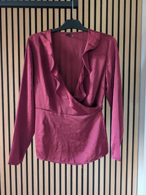 Vinröd omlottblus med volang - Snygg vinröd omlottblus med långa ärmar och djup v-ringning. Blusen har volangdetaljer längs ringningen och knyts i sidan för en feminin look. Materialet ser ut att vara ett mjukt tyg med lätt glans.