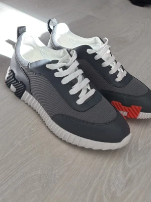 Grå sneakers med vita och orange detaljer - Snygga sneakers i grått med vita snören och sula. Skorna har coola detaljer i orange och svart på sulan samt dragflik bak. Ovandelen är i mesh och syntetmaterial för en modern look. Perfekta för dig som gillar stilrena men ändå unika sneakers.