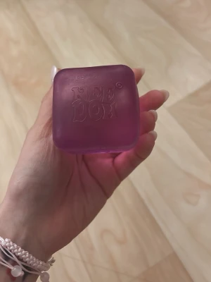Lila Nee Doh stressboll - En mjuk och klämbar lila stressboll från Nee Doh i fyrkantig form. Perfekt att ha i handen för att slappna av eller leka med. Tillverkad i ett mjukt, gummiaktigt material som är skönt att trycka på.