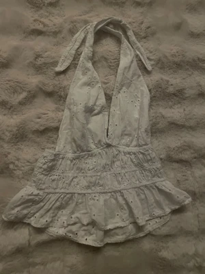 Vit broderad halterneck linnetopp - Söt vit halterneck topp med broderade detaljer och volangkant. Toppen knyts i nacken och har en omlott-look framtill. Perfekt för sommarens varma dagar och ger en romantisk vibe.