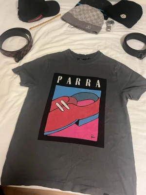 Grå Parra t-shirt med grafiskt tryck - Säljer en grå t-shirt från Parra med ett stort, färgglatt grafiskt tryck på bröstet. Trycket har motiv i rött, blått, rosa och vitt med texten 'PARRA' upptill. T-shirten har korta ärmar och en klassisk passform.