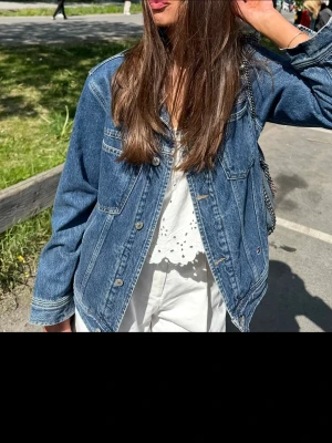 Blå Tommy Jeans jeansjacka - Säljer en klassisk blå oversize jeansjacka från Tommy Jeans med stort logopatch i rött, vitt och blått på ryggen. Jackan har knappstängning framtill, två bröstfickor med lock och en avslappnad passform. Perfekt för dig som gillar streetstyle och tidlös denim.