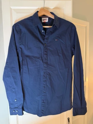 Tommy Hilfiger skjorta - Snygg marinblå skjorta från Tommy Jeans i slim fit-modell. Skjortan har långa ärmar, klassisk krage och knappar framtill. Tillverkad i bomull och har en liten logga broderad på bröstet. Perfekt för en clean och stilren look.