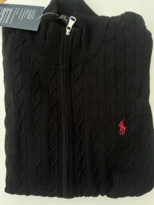 Svart kabelstickad tröja Polo Ralph Lauren - Kabelstickad svart tröja med dragkedja från Polo Ralph Lauren. Tröjan har hög krage och den klassiska röda logotypen broderad på bröstet. Tillverkad i mjukt material och har ribbade muddar vid hals och ärmslut.