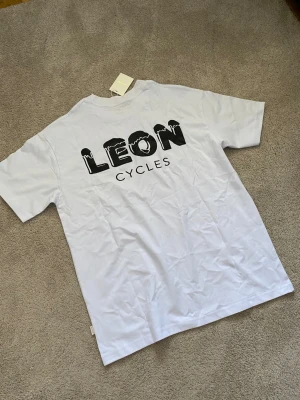 Vit Aime Leon Dore LEON CYCLES t-shirt - Säljer en vit t-shirt med tryck från Aime Leon Dore. Framsidan har en liten svart logga med vingar och cykelmotiv, medan baksidan har en stor text med LEON CYCLES i svart. Klassisk passform och rund hals. Perfekt för dig som gillar cykelstil.