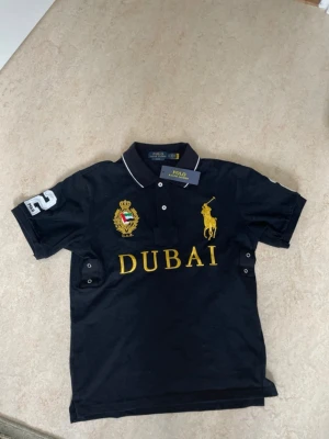 Svart piké från Polo Ralph Lauren - Svart pikétröja från Polo Ralph Lauren med gul brodyr av logga och texten DUBAI på bröstet. Har även ett emblem med UAE-flagga och siffran 2 på ärmen. Klassisk krage med vita detaljer och knappar framtill.