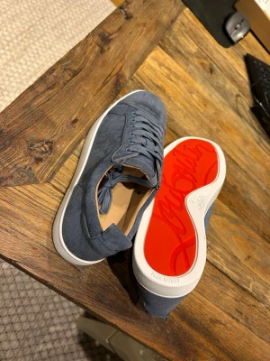 Loubs Blå mockasneakers - Snygga blå sneakers i mocka med klassisk låg profil och vita sulor. Skorna har en ikonisk röd undersida och matchande blå skosnören. Perfekta för dig som vill ha en stilren men ändå unik look.